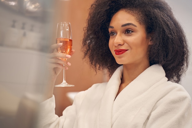 Mujer de moda de pelo rizado mirando hacia otro lado mientras se relaja con una copa de vino champán