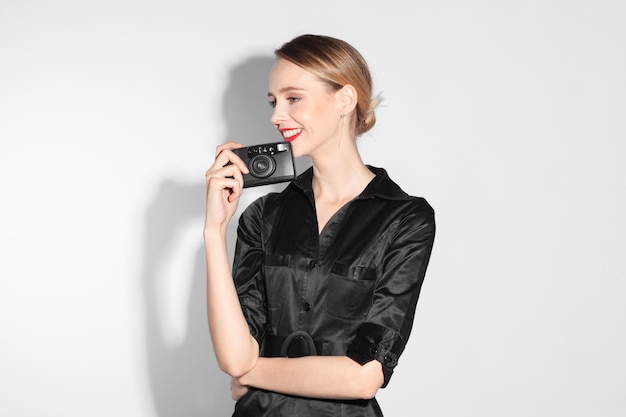 Mujer de moda sonriente en vestido negro con labios rojos con cámara vintage aislada sobre fondo blanco