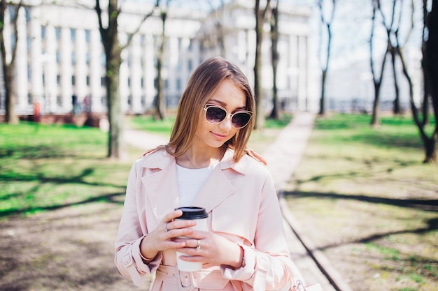 Mujer de moda con teléfono y café en la ciudad. Mujer de moda en gafas de sol y chaqueta rosa al aire libre