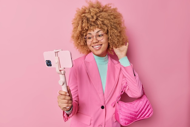 Foto una mujer de moda vestida con ropa formal sostiene un teléfono inteligente en un palo de selfie y toma una foto con una bolsa de damas que sonríe felizmente aislada sobre un fondo rosa una dama elegante se prepara para una reunión formal