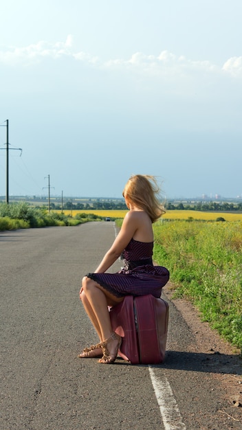 Mujer de moda en un vestido de verano y tacones de aguja de espaldas a la cámara haciendo autostop en la carretera en el campo