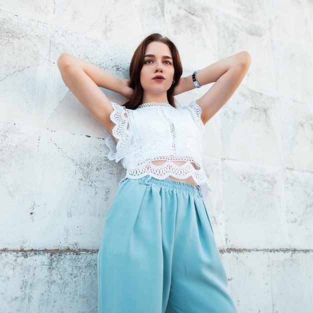 Mujer modelo joven bastante moderna en una blusa de encaje de elegancia en pantalones azules de moda posa cerca de una pared blanca vintage en la ciudad.