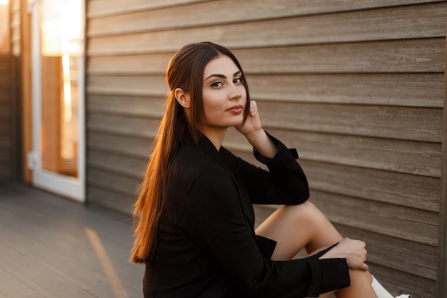 Mujer modelo muy elegante en un elegante abrigo negro se sienta cerca de una pared de madera al atardecer