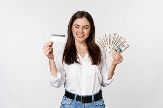 Mujer moderna feliz con dinero y tarjeta de crédito en las manos, sonriendo complacida, de pie sobre fondo blanco. copia espacio