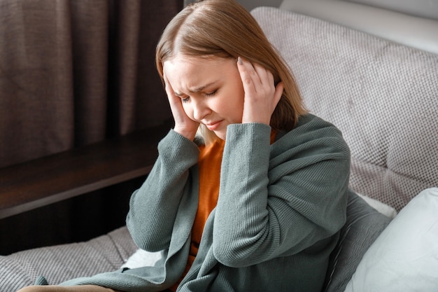Mujer molesta con migraña severa sostiene las sienes con las manos. La mujer tiene fiebre y dolor de cabeza sentada en su casa solo en el sofá.