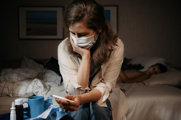 Foto mujer molesta y preocupada sentada en la cama y mirando el termómetro niño pequeño acostado en la cama