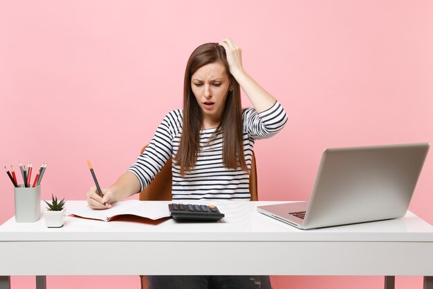 Mujer molesta que se aferra a la cabeza usando la calculadora escribiendo notas con cálculos sentarse y trabajar en la oficina con un ordenador portátil aislado sobre fondo rosa pastel. Concepto de carrera empresarial de logro. Copie el espacio.