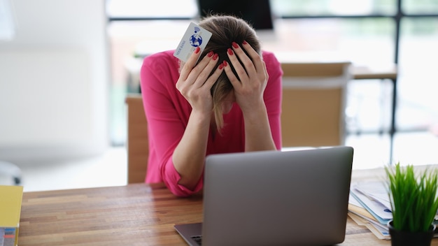 Mujer molesta sentada en una laptop con tarjeta plástica de crédito y sosteniendo su cabeza