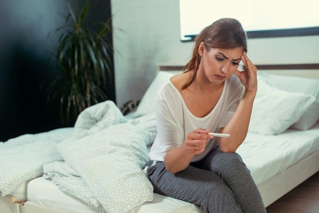 Mujer molesta sentada triste en una cama blanca con una prueba de embarazo negativa