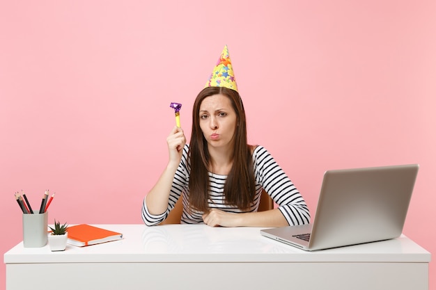 Mujer molesta en sombrero de fiesta de cumpleaños con pipa de juego celebrando solo ofenderse mientras se sienta a trabajar en el escritorio blanco con computadora portátil pc