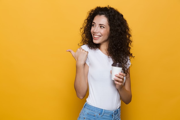 Mujer morena de 20 años con cabello rizado sonriendo y sosteniendo café para llevar en un vaso de papel aislado en amarillo