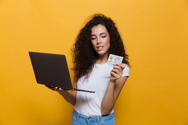 Foto mujer morena de 20 años vistiendo ropa casual con portátil negro y tarjeta de crédito aislado en amarillo