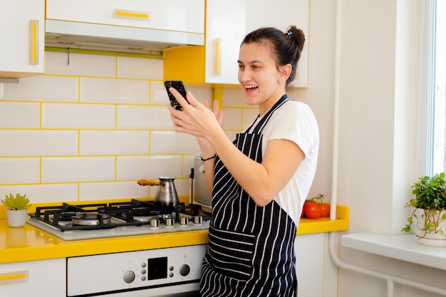 Mujer morena de 30 a 35 años con delantal blanco y negro usando un smartphone para videollamadas y haciendo gestos en la cocina. Concepto concepto de comunicación remota, cuarentena.