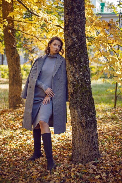 Una mujer morena con un abrigo gris y botas está de pie junto a un árbol en otoño entre los árboles