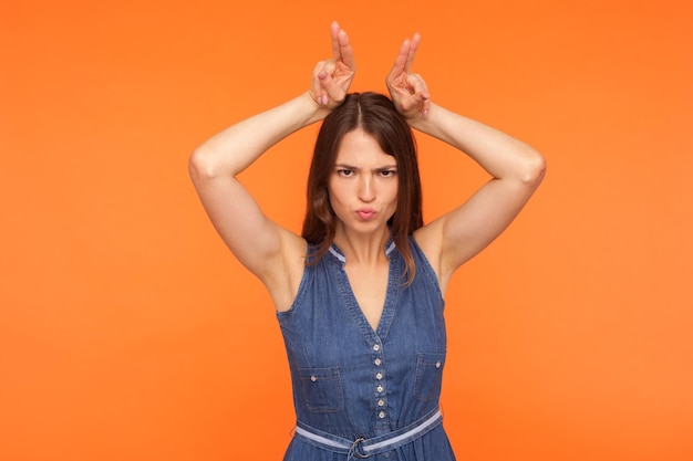 Mujer morena acosadora enojada con vestido de mezclilla que muestra cuernos de toro con los dedos en la cabeza, gesto de asta, amenazando con una vista hostil, lista para atacar. tiro de estudio interior aislado sobre fondo naranja