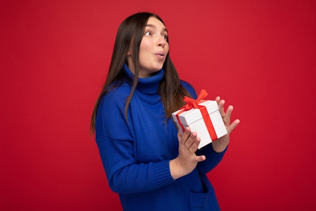 mujer morena aislada sobre pared roja vistiendo un suéter azul casual sosteniendo una caja de regalo blanca y mirando hacia el lado.