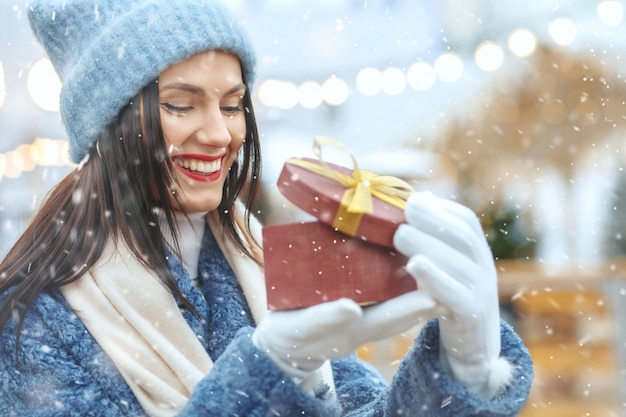 Mujer morena alegre en abrigo de invierno sosteniendo una caja de regalo en la feria de Navidad durante las nevadas. Espacio para texto