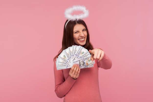 Mujer morena alegre con halo sobre su cabeza sosteniendo billetes de dólar apuntándote con el dedo con una sonrisa dentuda ganadora de la lotería éxito Estudio interior aislado en fondo rosa