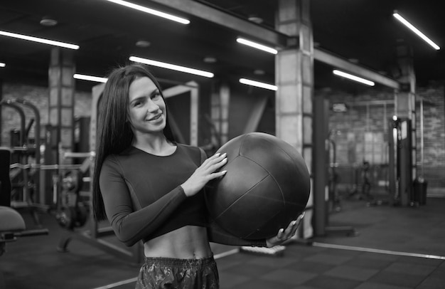 Mujer morena alegre en ropa deportiva sosteniendo balón medicinal en la mano y posando a la cámara.