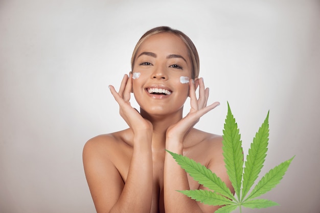 Mujer morena aplicando crema facial con CBD a base de extracto de cannabis. Aislado sobre fondo gris