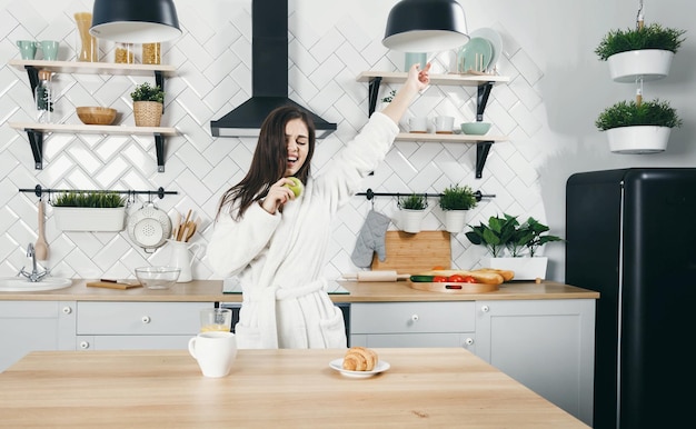 Mujer morena en bata blanca canta una canción a la manzana como un micrófono en la cocina