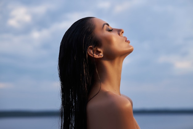 Mujer morena de belleza en un vestido negro posa en un lago de mar sobre un fondo de cielo azul. Mujer sexy de pelo largo y maquillaje de belleza hermosa en su rostro