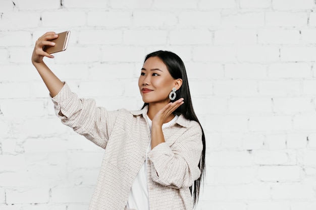 Mujer morena bronceada en elegante cárdigan beige toma selfie sobre fondo de pared de ladrillo blanco Atractiva dama sostiene teléfono y sonríe