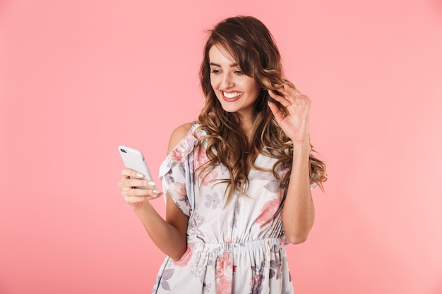 Mujer morena con cabello largo sosteniendo y usando un teléfono inteligente, aislado en rosa