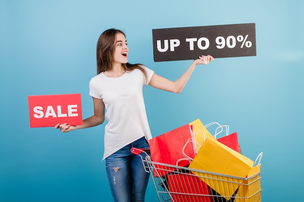 Foto mujer morena con carretilla de compras llena de coloridas bolsas de papel rojo y amarillo y 90% cartel de venta aislado sobre azul