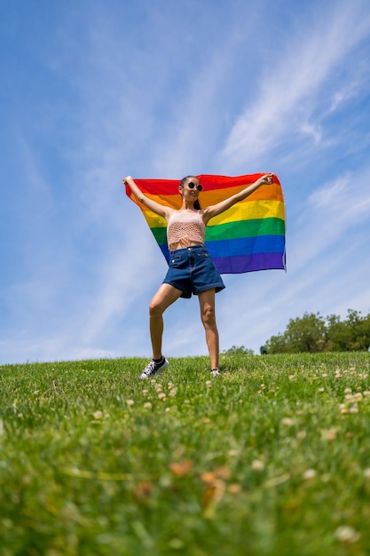 Mujer morena caucásica con una bandera lgbt del arco iris en el fondo de hierba y cielo azul