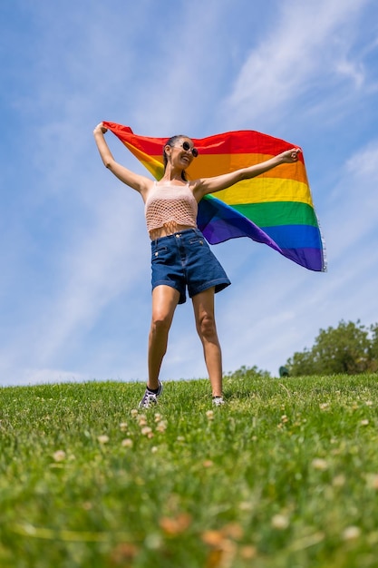 Mujer morena caucásica con una bandera lgbt de arco iris en la hierba y fondo de cielo azul ondeando la bandera