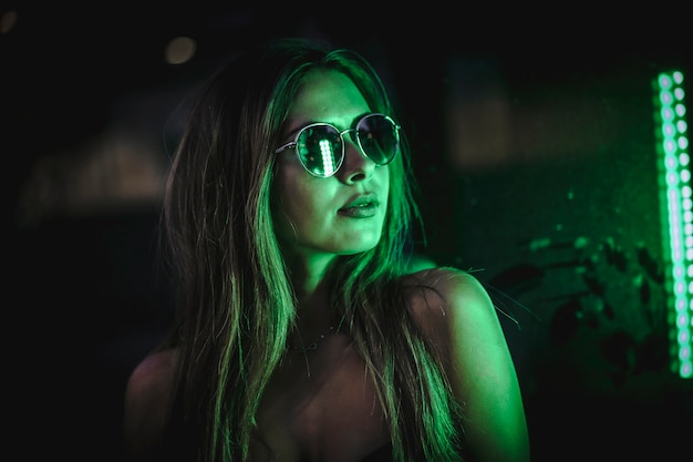 Mujer morena caucásica con gafas de sol mirando un leds verde claro. Fotografía urbana nocturna