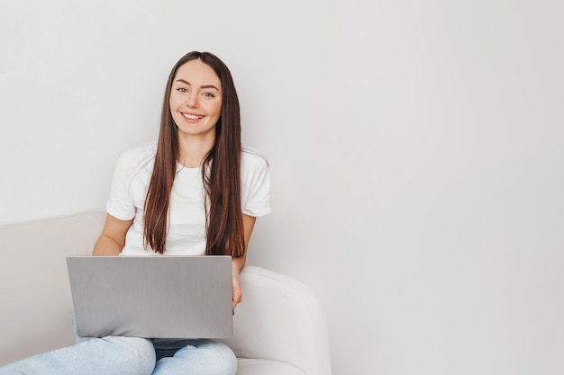 Mujer morena caucásica joven sonriendo a la cámara y sentada con la computadora portátil en el sofá