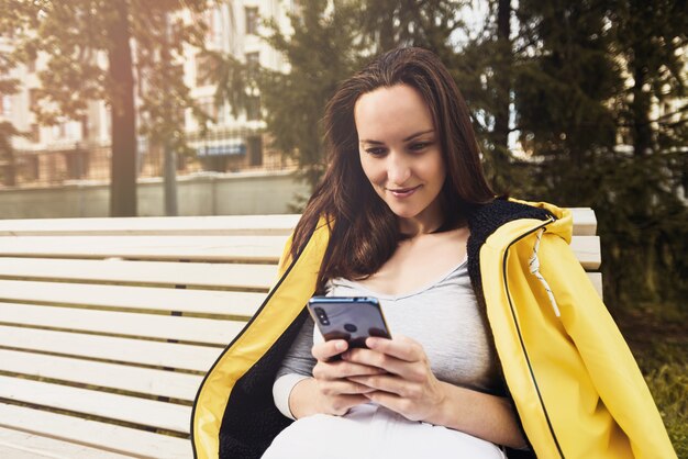 Mujer morena de chaqueta amarilla con smartphone en sus manos sentado en un banco