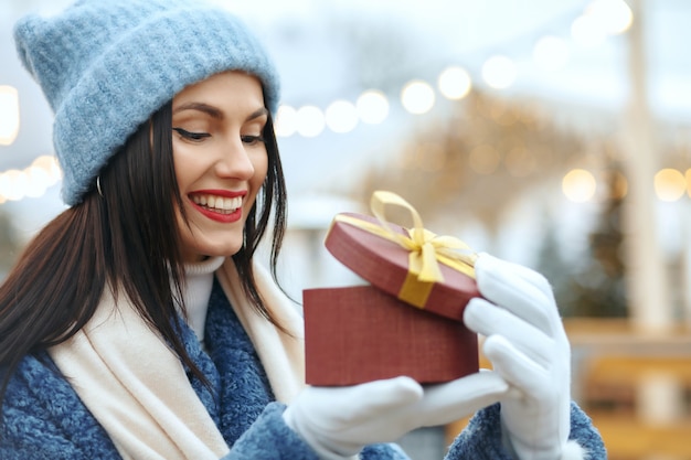 Mujer morena complacida en abrigo de invierno sosteniendo una caja de regalo en la feria de Navidad. Espacio para texto