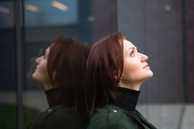 Una mujer morena con un corte de pelo bob está de espaldas a la pared espejada, se refleja y se ve triste. El tema de la soledad