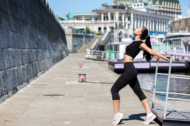 Mujer morena delgada con ropa deportiva que se extiende en la ciudad, cerca del río. Espacio para texto