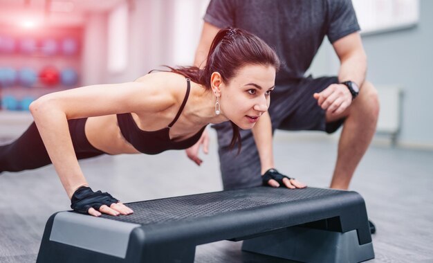 Mujer morena deportiva haciendo flexiones desde el soporte de plástico y un entrenador en el club deportivo Ejercicios de modelo de fitness joven en el gimnasio Primer plano