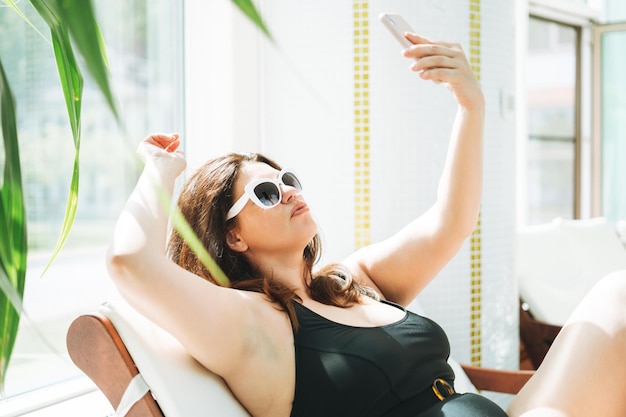 Mujer morena elegante cuerpo positivo de talla grande en traje de baño negro y gafas de sol tomando selfie en el teléfono móvil en la tumbona en el spa