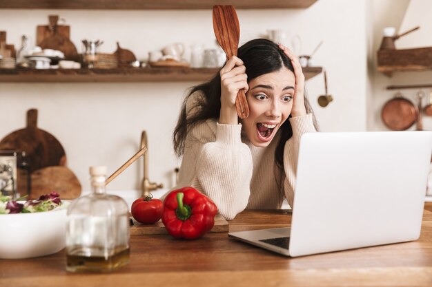 Mujer morena emocional con ordenador portátil mientras cocina ensalada verde saludable con verduras en la elegante cocina de casa