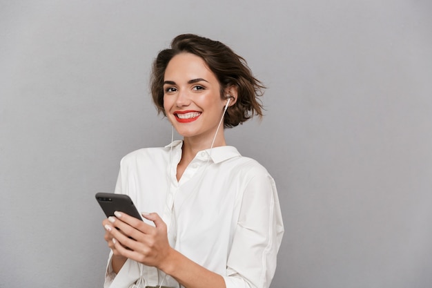 Mujer morena escuchando música en el teléfono inteligente a través de auriculares, aislado sobre una pared gris