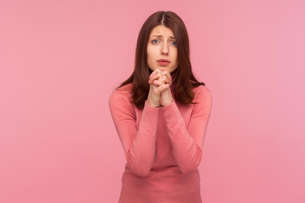 Mujer morena esperanzada con suéter rosa tomándose de la mano en oración, pidiendo perdón mirando a la cámara con respeto, necesita ayuda. Disparo de estudio interior aislado sobre fondo rosa