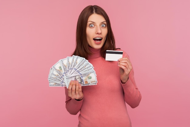 Mujer morena extremadamente sorprendida mostrando tarjeta de crédito y fanático de los billetes en dólares sorprendida con una alta tasa de interés en el depósito Foto de estudio interior aislada sobre fondo rosa