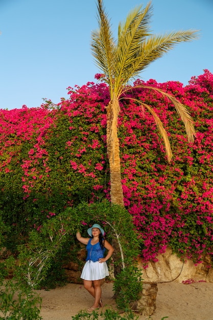 Una mujer morena con una falda blanca y un sombrero para el sol se encuentra junto a las plantas verdes en flor cerca del arco