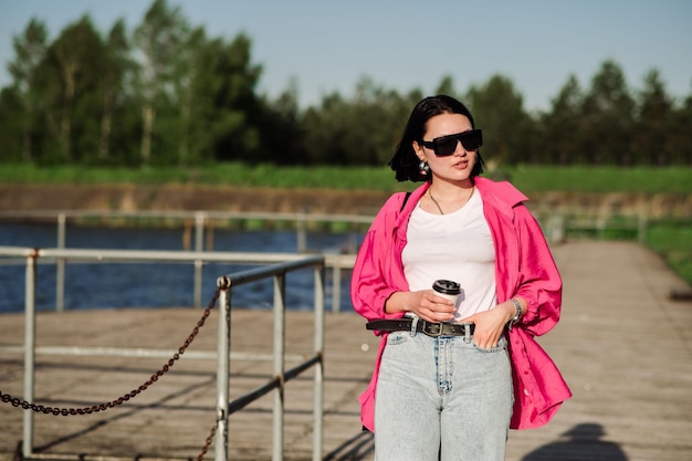 Mujer morena feliz con gafas de sol caminando cerca del lago en un muelle de madera en un día soleado