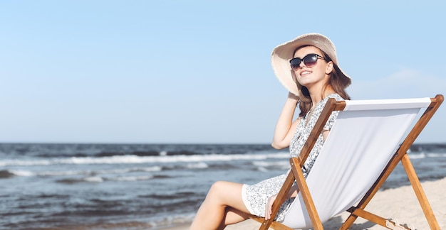 Mujer morena feliz con gafas de sol y sombrero relajándose en una tumbona de madera en la playa del océano.