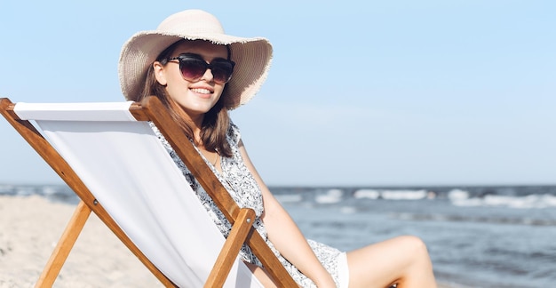 Mujer morena feliz con gafas de sol y sombrero relajándose en una tumbona de madera en la playa del océano