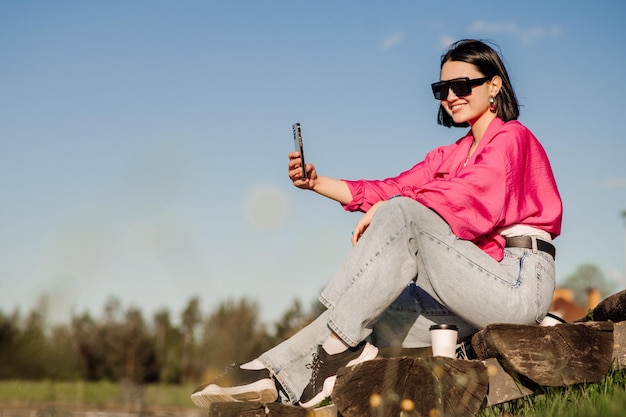 Una mujer morena feliz con gafas de sol se toma un selfie usando un celular sobre el cielo azul al aire libre en el parque