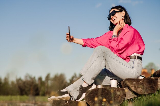 Una mujer morena feliz con gafas de sol se toma un selfie usando un celular sobre el cielo azul al aire libre en el parque
