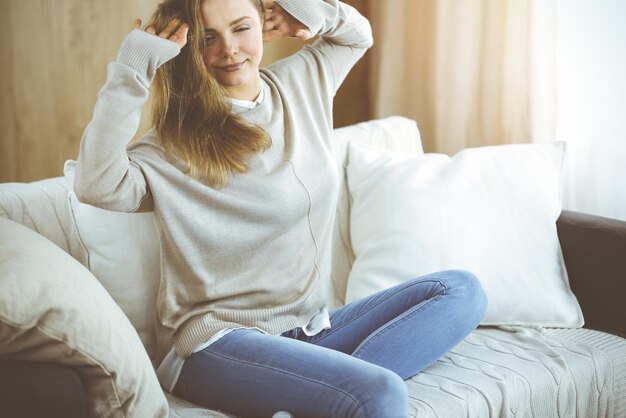 Mujer morena feliz puso las manos detrás de la cabeza sentada apoyada en el sofá, mujer europea estirándose y disfrutando de un fin de semana perezoso o de vacaciones en una acogedora sala de estar.